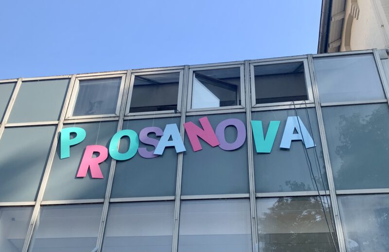Aus den Fenstern eines Schulgebäudes hängen große, bunte Pappbuchstaben, die das Wort Prosanova ergeben.