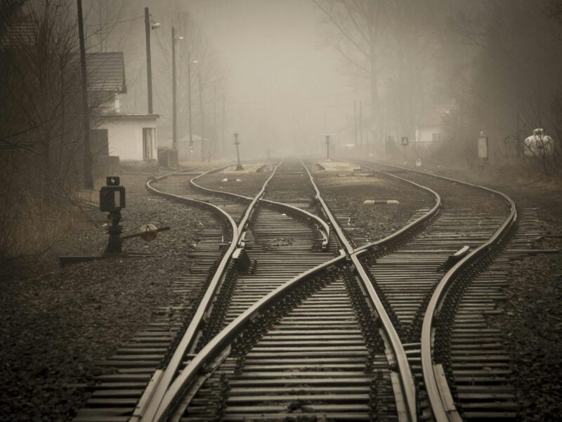 Ein Gleis führt durch Weichen in mehrere Richtungen. Links im Bild ist ein Haus zu sehen und im Hintergrund kahle Bäume, die im Nebel verschwinden. Das Bild ist in schwarz-weiß.